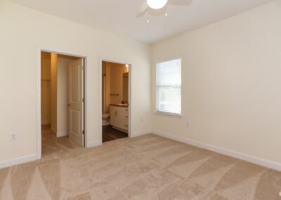 An empty bedroom with freshly-vacuumed beige carpet, a white ceiling fan, and an en-suite bathroom
