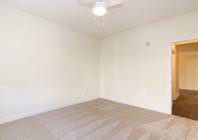 An empty bedroom with off-white walls, freshly-vacuumed beige carpet, an a white ceiling fan.