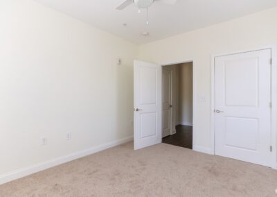 An empty bedroom with off-white walls, beige carpet, and a white ceiling fan.