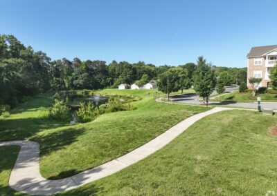 A lush green park with a concrete path winding through it. There is a large pond with a fountain near the path.