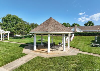 An open-air gazebo with some metal patio furniture in the middle of a grassy area with concrete paths leading to the gazebo.